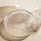 The Mini Lily Diamond Bracelet, featuring small sparkling stones, is displayed in a round, clear ribbed jewelry case on a beige surface—ideal for fans of minimal bracelet styles.