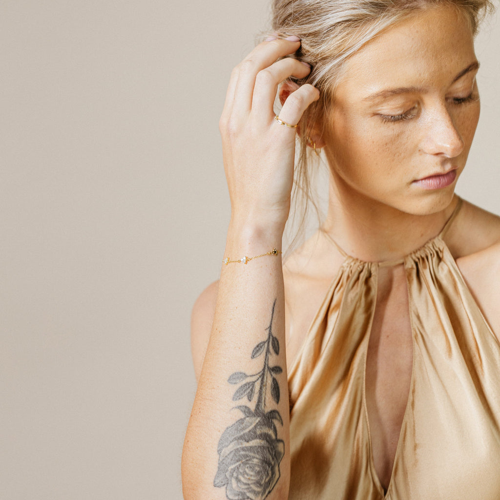 A woman in a gold satin top with a rose tattoo on her arm wears gold jewelry and the Mini Lily Diamond Bracelet, gazing down thoughtfully.