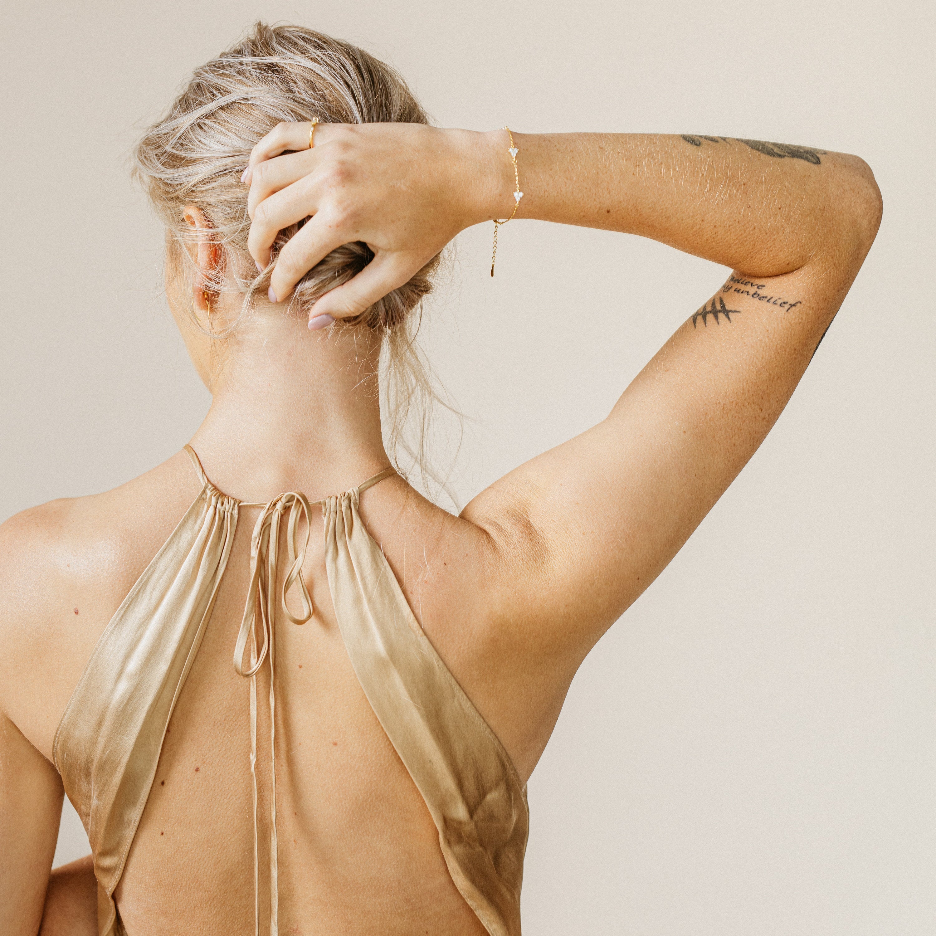 A woman in a gold halter dress, back to the camera and hand in her hair, displays her tattoos and the Mini Lily Diamond Bracelet, adding sparkle to her jewelry collection.