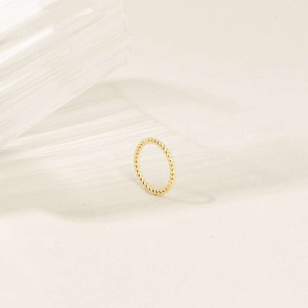 The Lea Twisted Ring, featuring polished bands, rests on a light surface next to a clear, textured glass object.