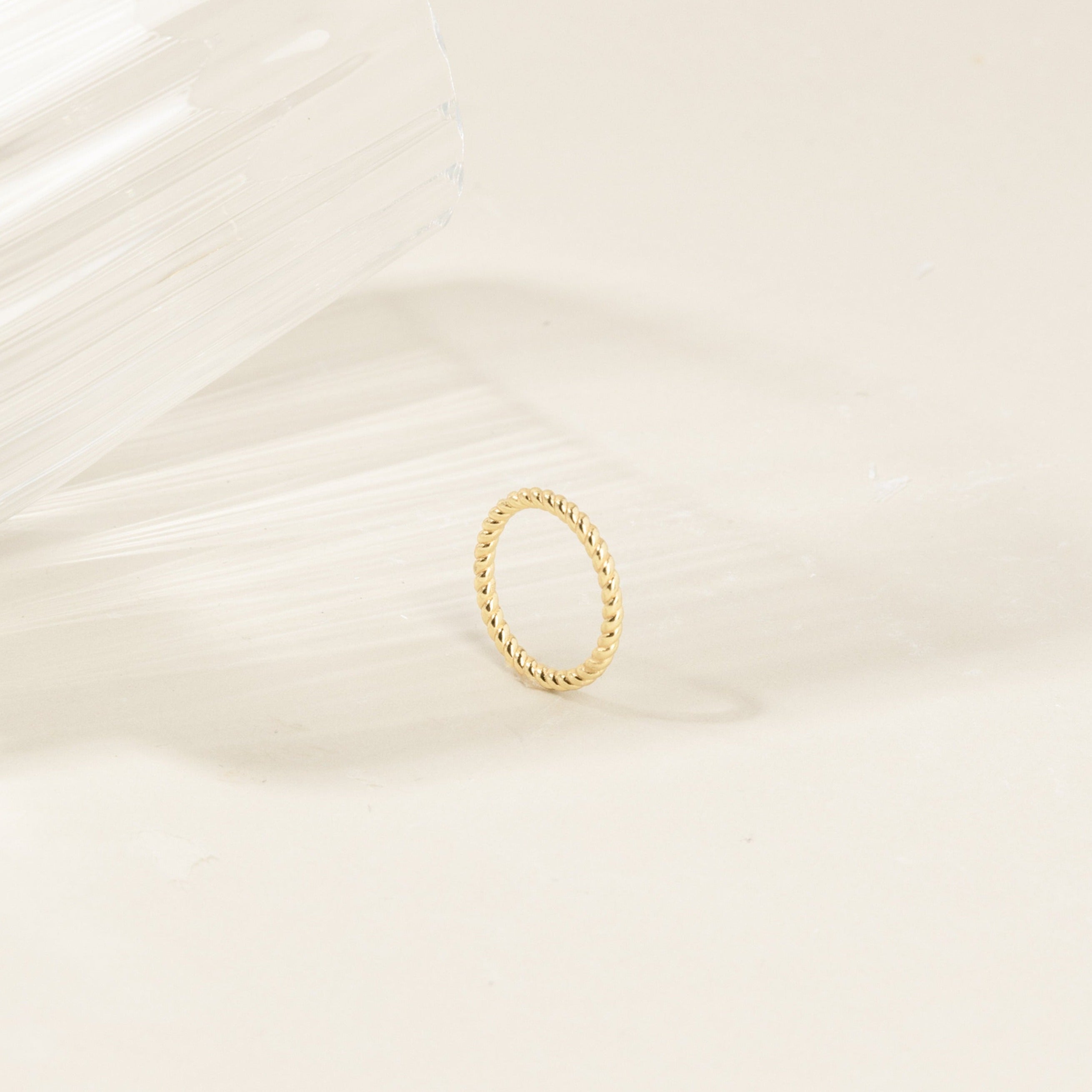 The Lea Twisted Ring, featuring polished bands, rests on a light surface next to a clear, textured glass object.