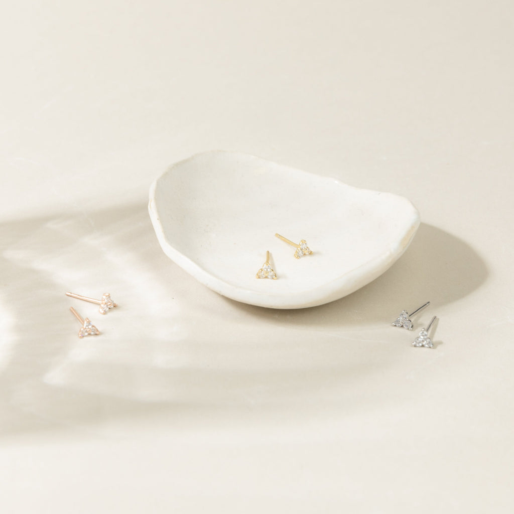 Three pairs of Mini Lily Studs—triangle-shaped earrings in gold, rose gold, and silver—ideal for cartilage piercings or daily wear, shown near a white ceramic dish.