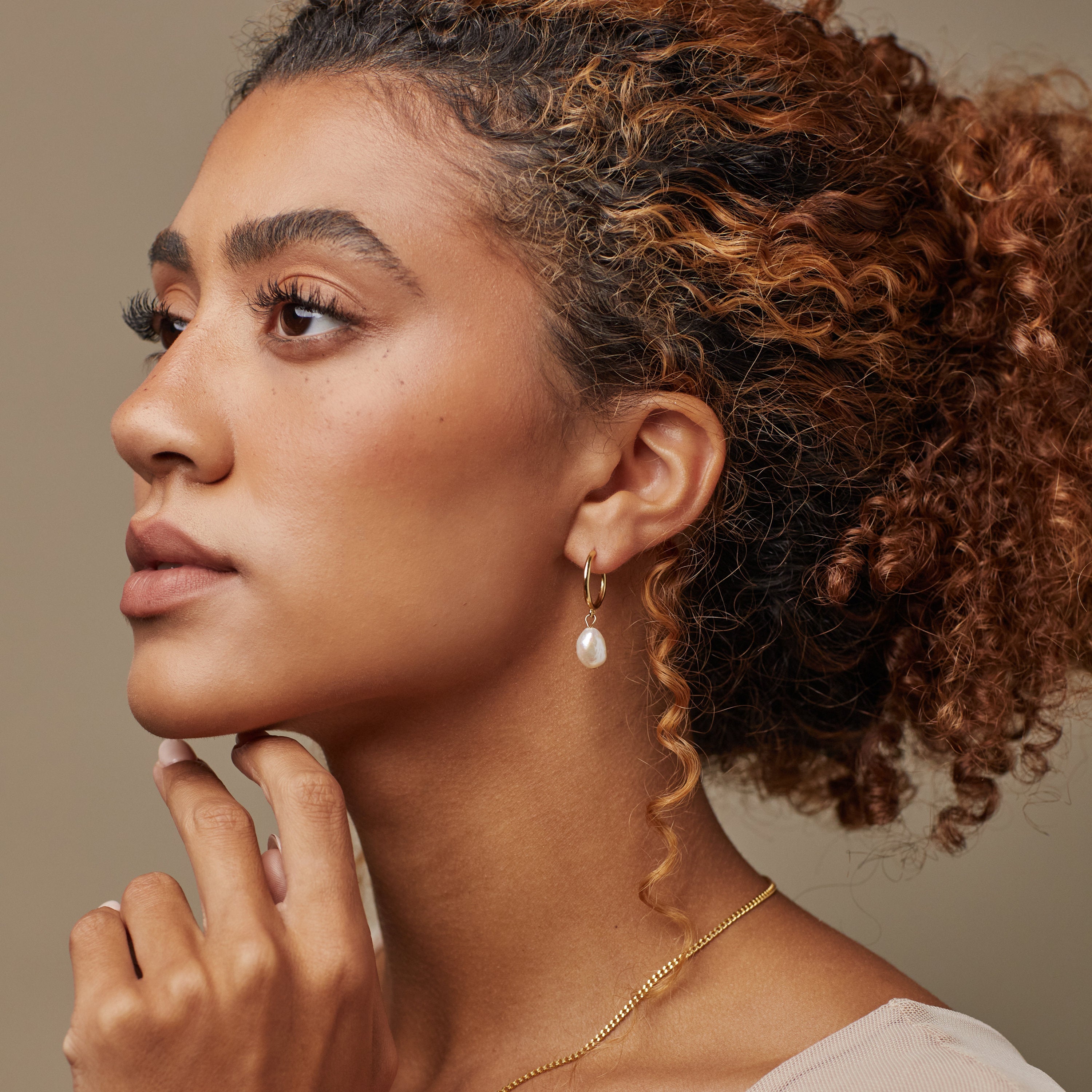 Woman with curly hair, wearing a gold necklace and Margot Pearl Hoops, looks thoughtfully to the side.