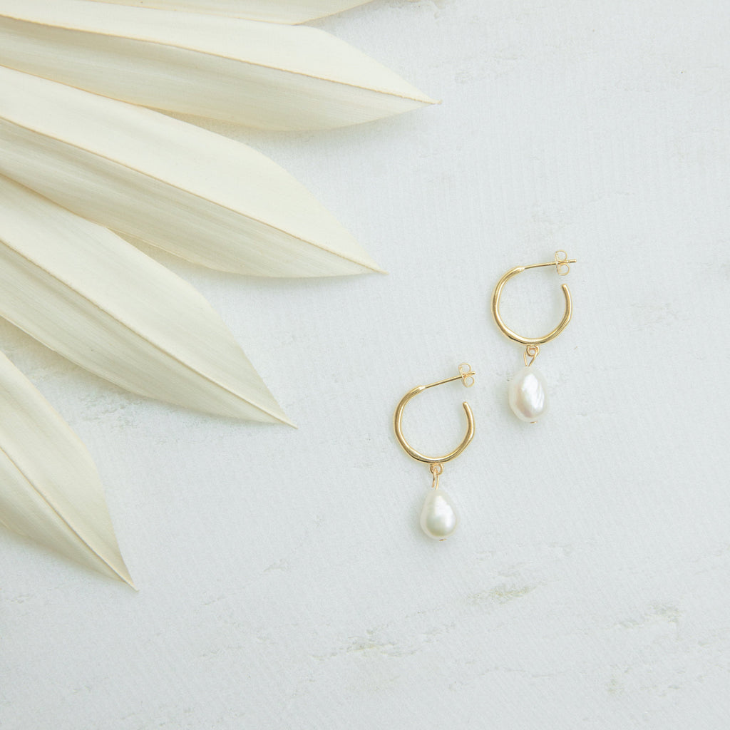 Margot Pearl Hoops, featuring gold hoops with drop pearl accents, are displayed on a light surface beside pale dried palm leaves.