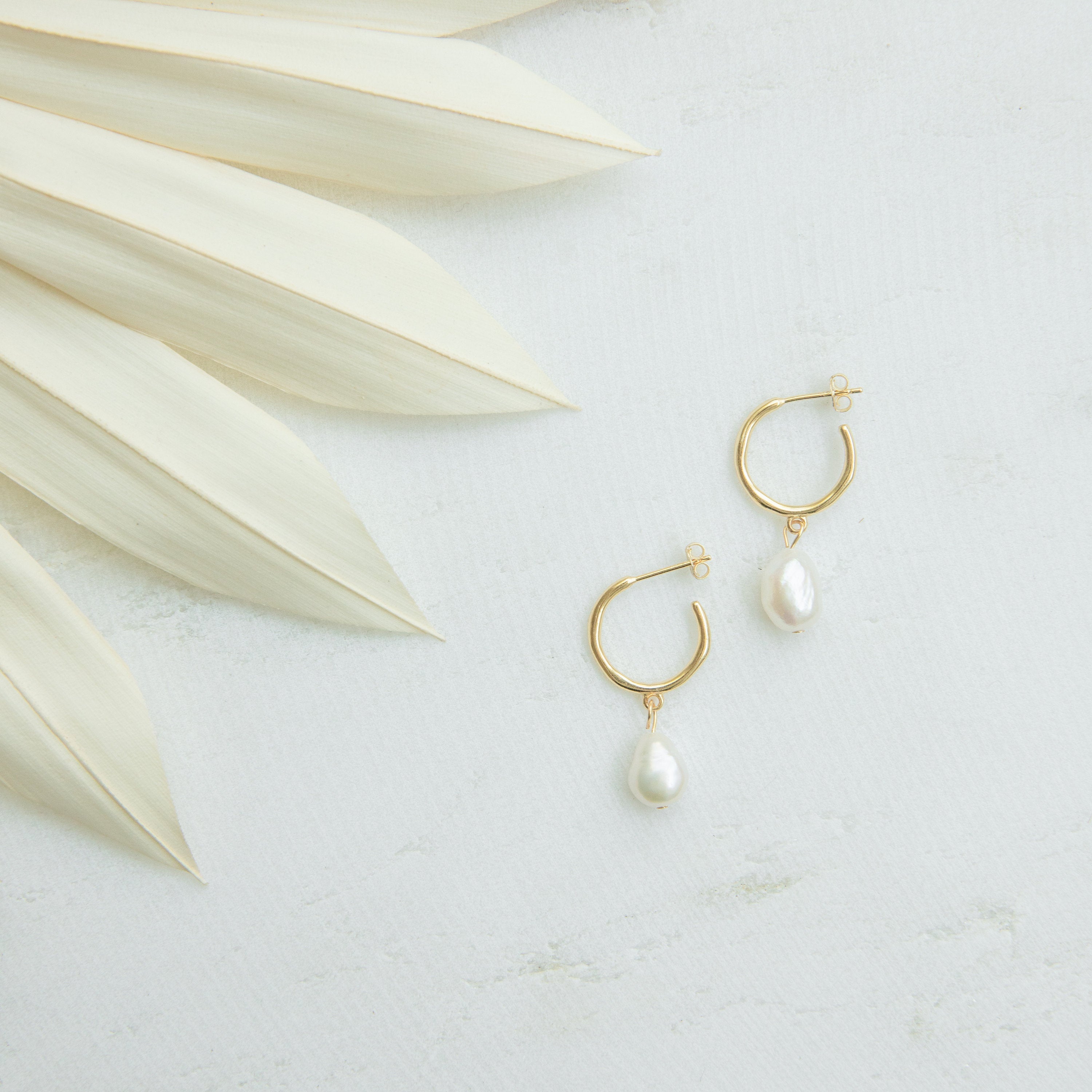 Margot Pearl Hoops, featuring gold hoops with drop pearl accents, are displayed on a light surface beside pale dried palm leaves.
