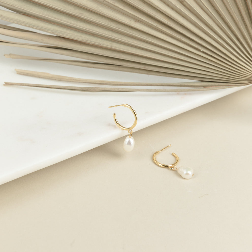 Margot Pearl Hoops—gold hoop earrings with delicate drop pearls—are displayed on a marble and beige surface, accented by a decorative palm leaf in the background.