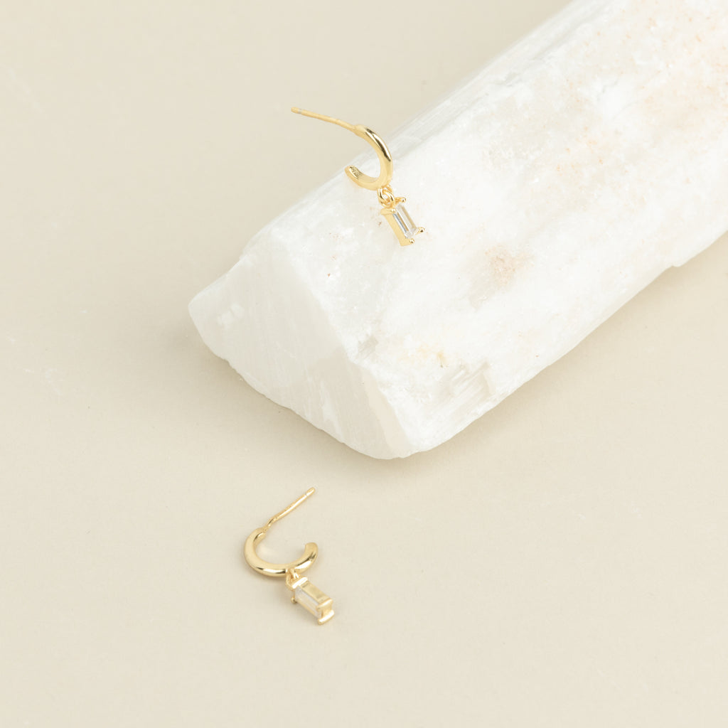 Two Lys Baguette Hoops, each featuring rectangular clear gemstones, display classic elegance—one rests on a white stone, the other on a beige surface.
