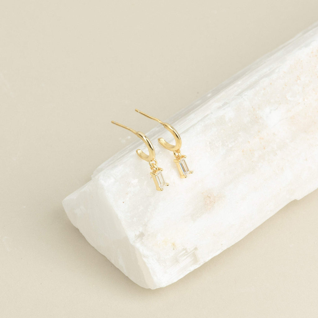 Lys Baguette Hoops feature gold earrings with rectangular crystals, capturing classic elegance and displayed on a white stone slab against a beige background.