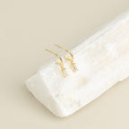 Lys Baguette Hoops feature gold earrings with rectangular crystals, capturing classic elegance and displayed on a white stone slab against a beige background.