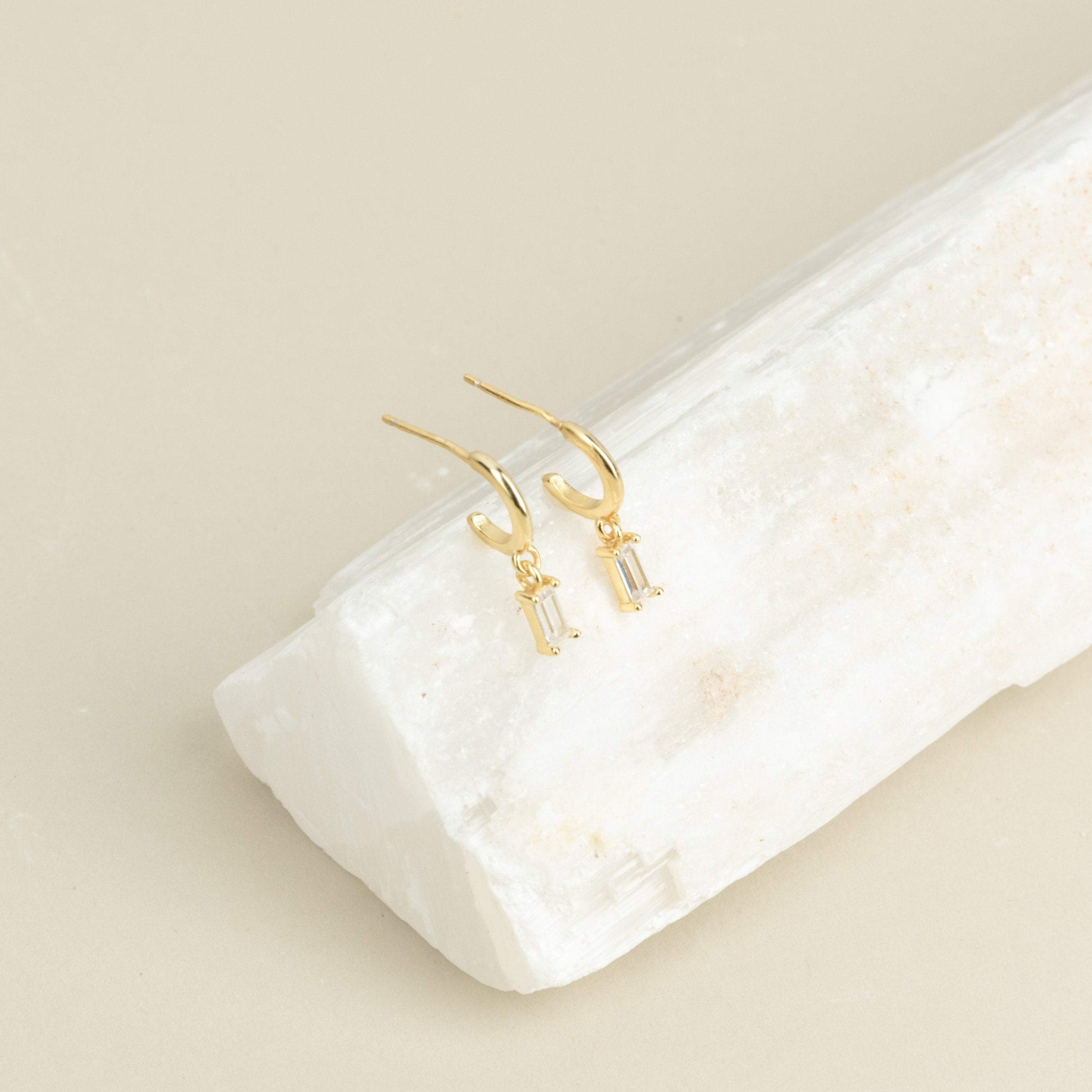 Lys Baguette Hoops feature gold earrings with rectangular crystals, capturing classic elegance and displayed on a white stone slab against a beige background.