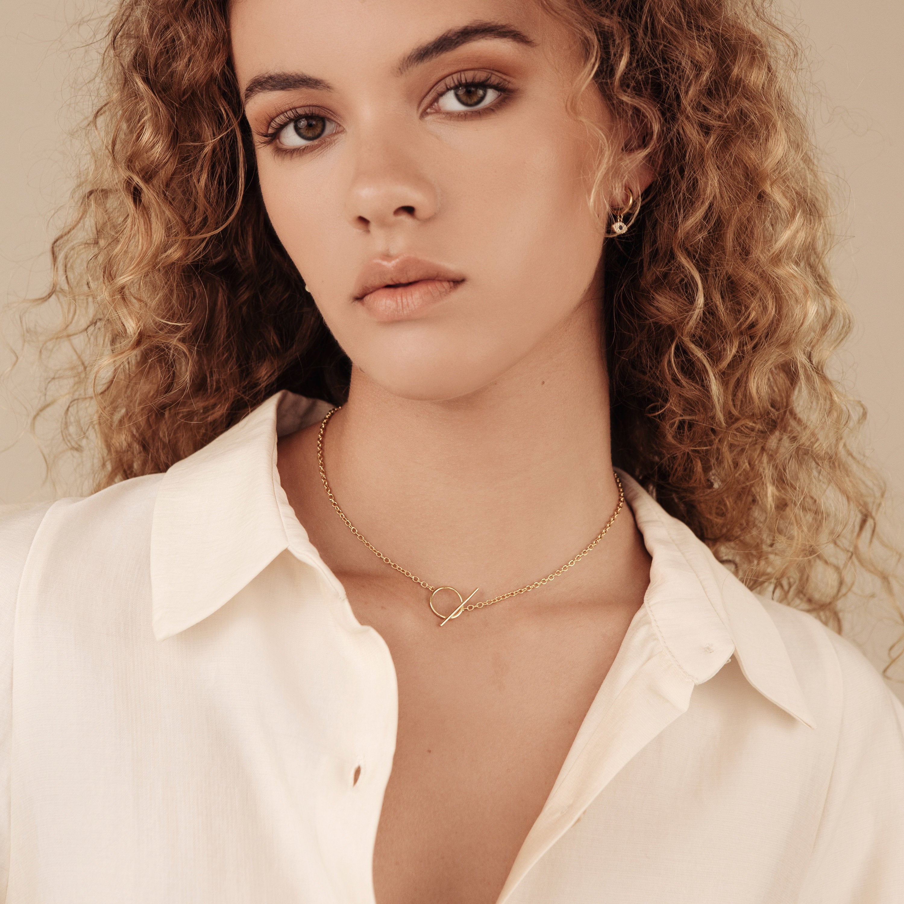 Woman with curly hair wears a cream blouse and minimal jewelry, featuring the Skyy Toggle Necklace in gold, as she looks at the camera with a neutral expression.