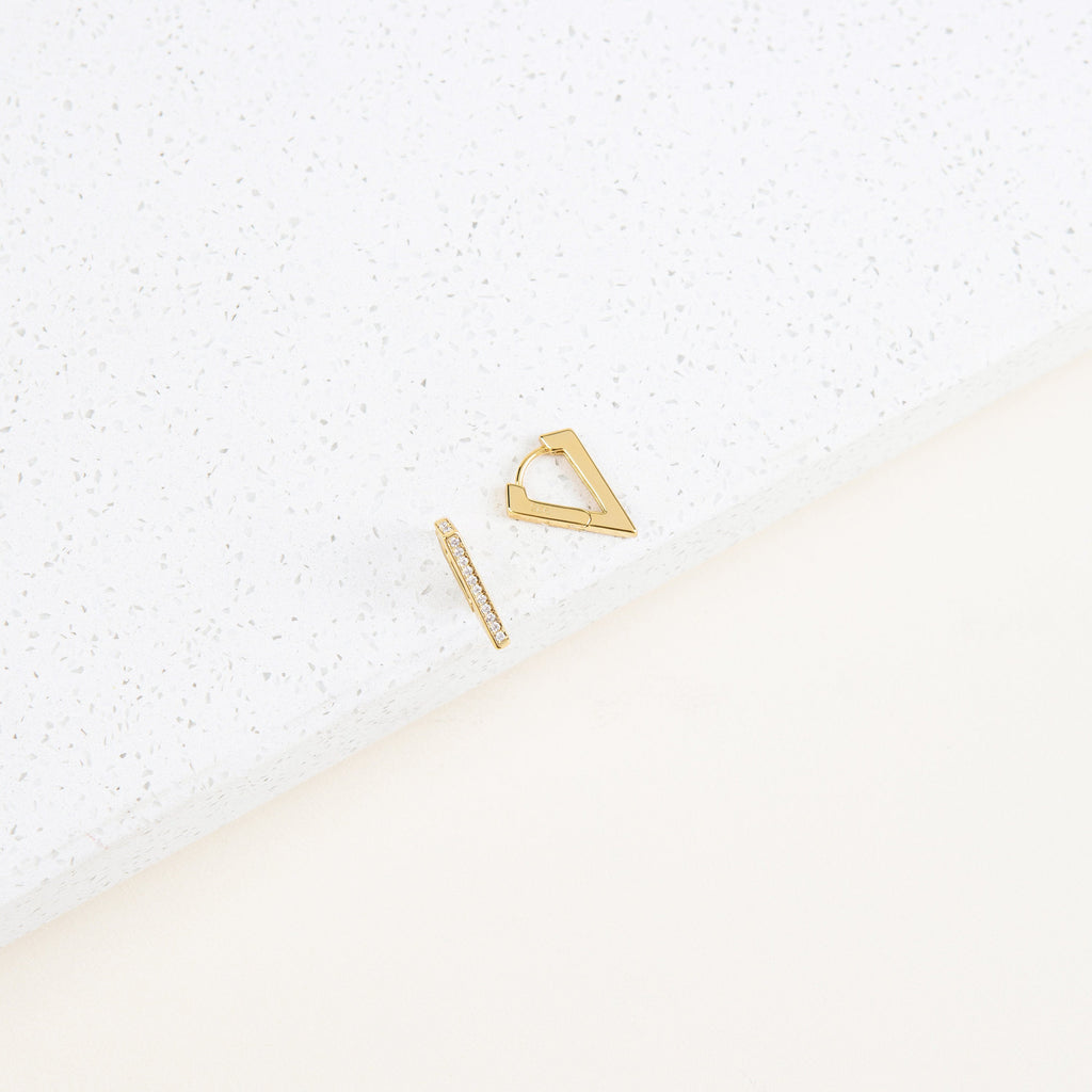 The Ezra Triangle Earrings—featuring one geometric triangle and one slim bar—are displayed on a white textured surface, highlighting their modern design and subtle pavé texture.