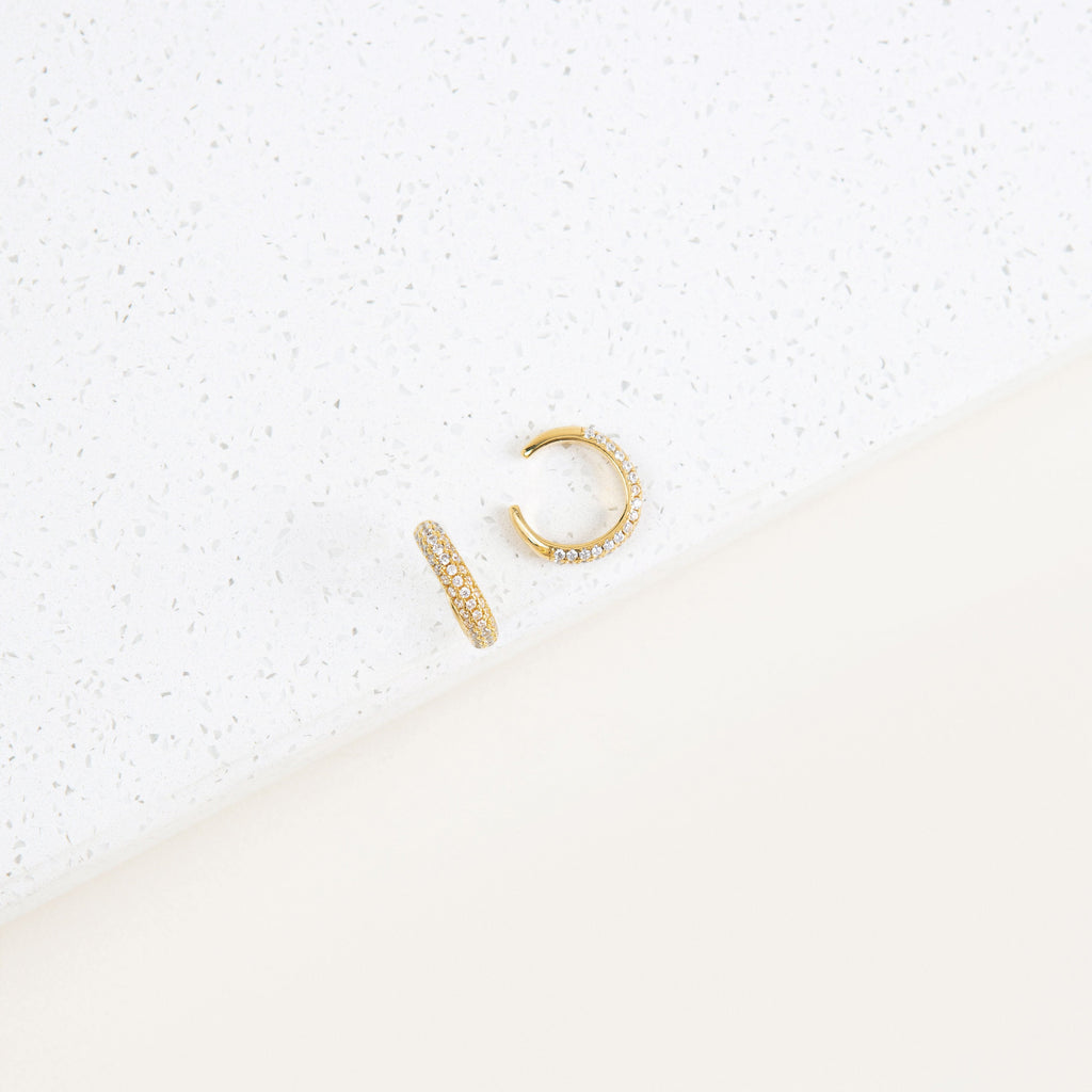 The Mia Pave Cuffs, gold hoop earrings adorned with small clear stones, sparkle beautifully on a white speckled surface.
