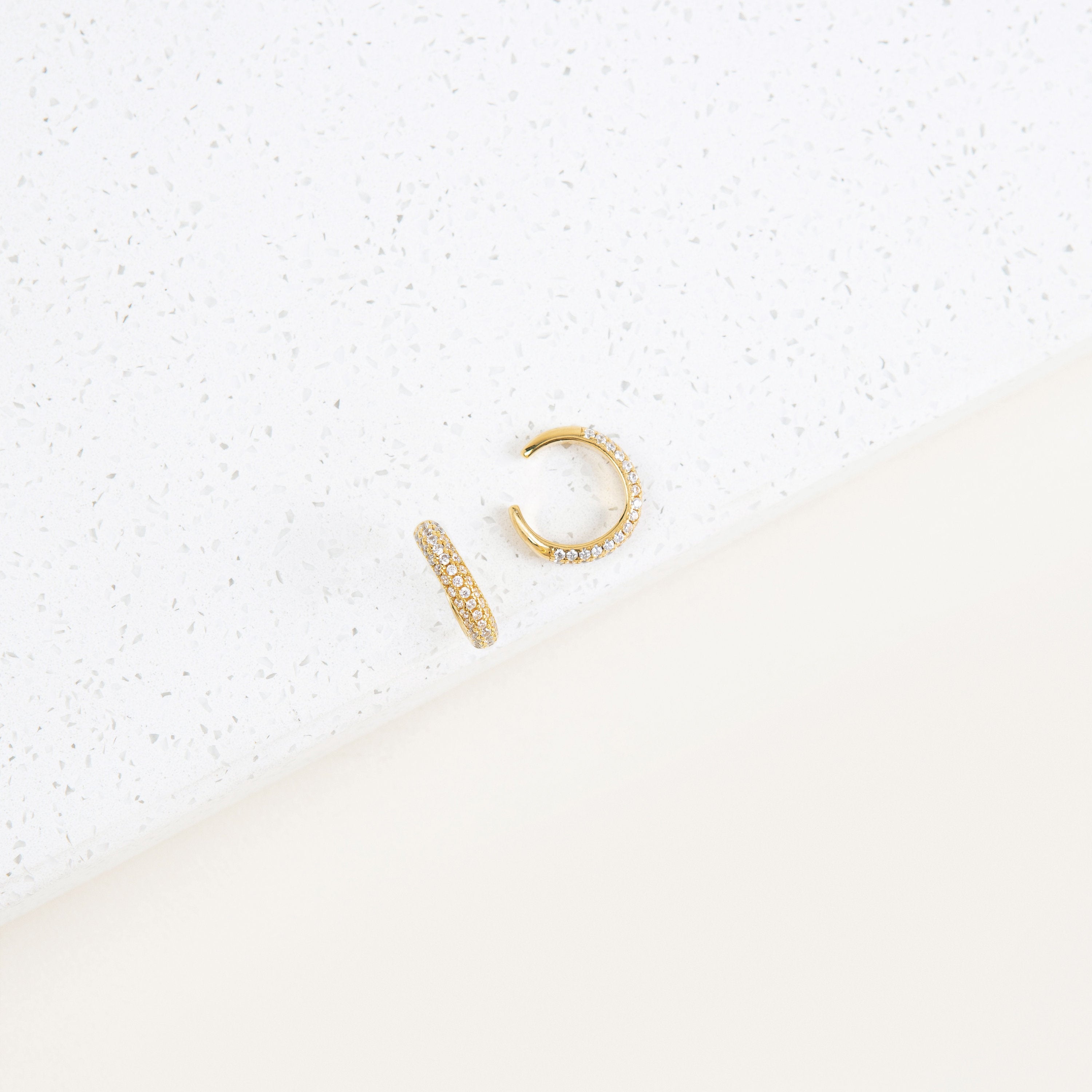 The Mia Pave Cuffs, gold hoop earrings adorned with small clear stones, sparkle beautifully on a white speckled surface.