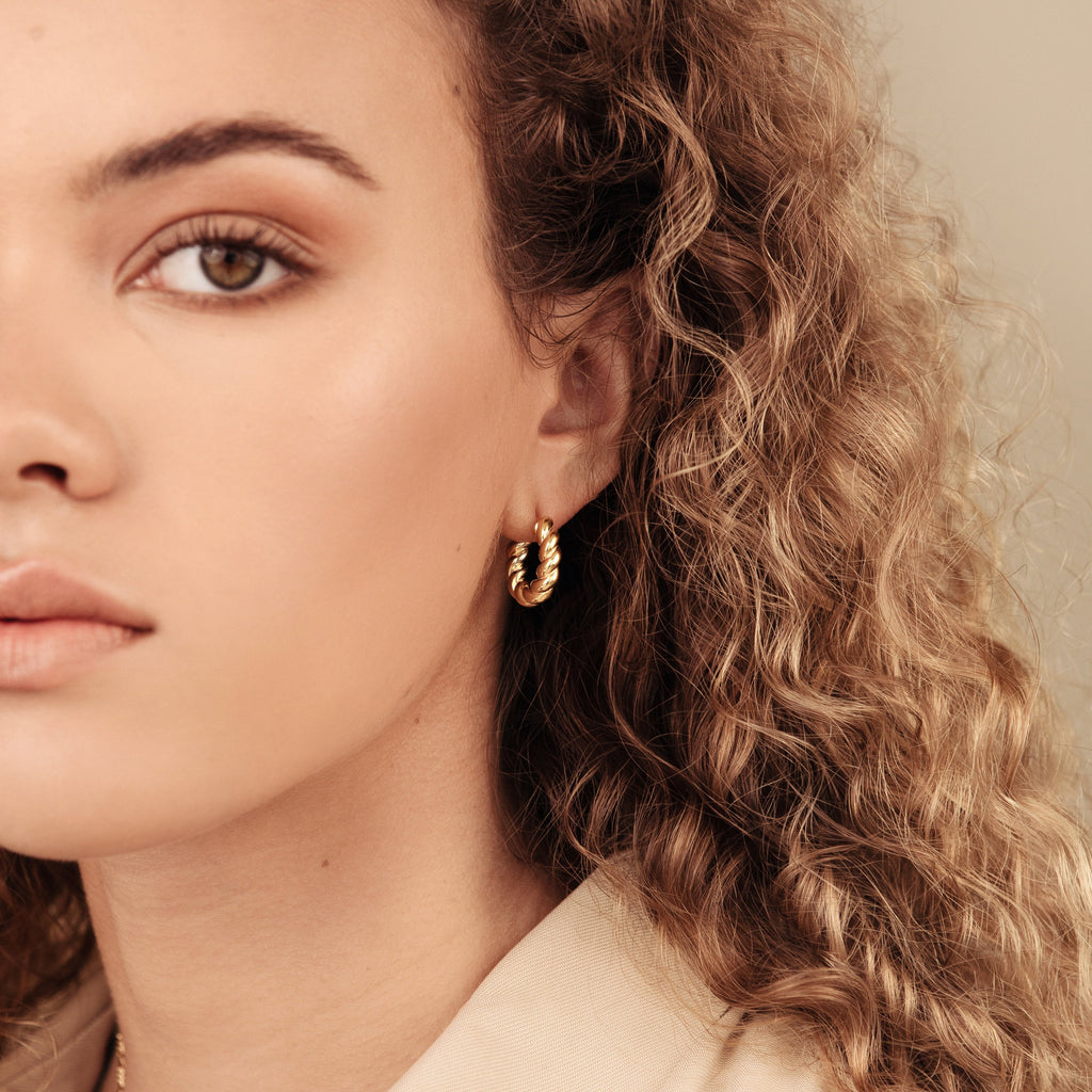 Close-up of a woman with curly hair wearing minimal jewelry: the Remi Twist Hoops, elegant gold earrings, while looking forward.