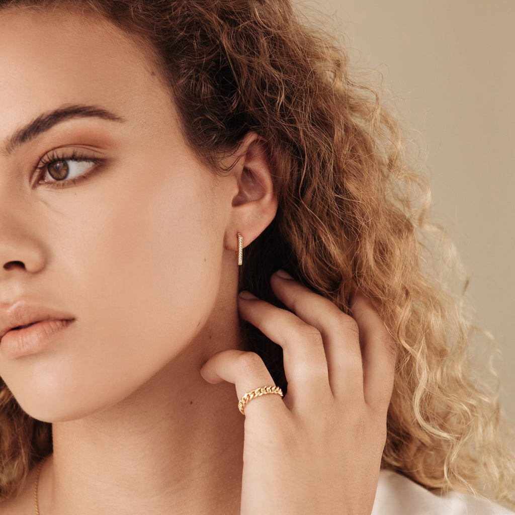 A woman with curly hair gently touches her ear, wearing a gold ring and the Ezra Triangle Earrings—modern geometric earrings with a pavé texture.