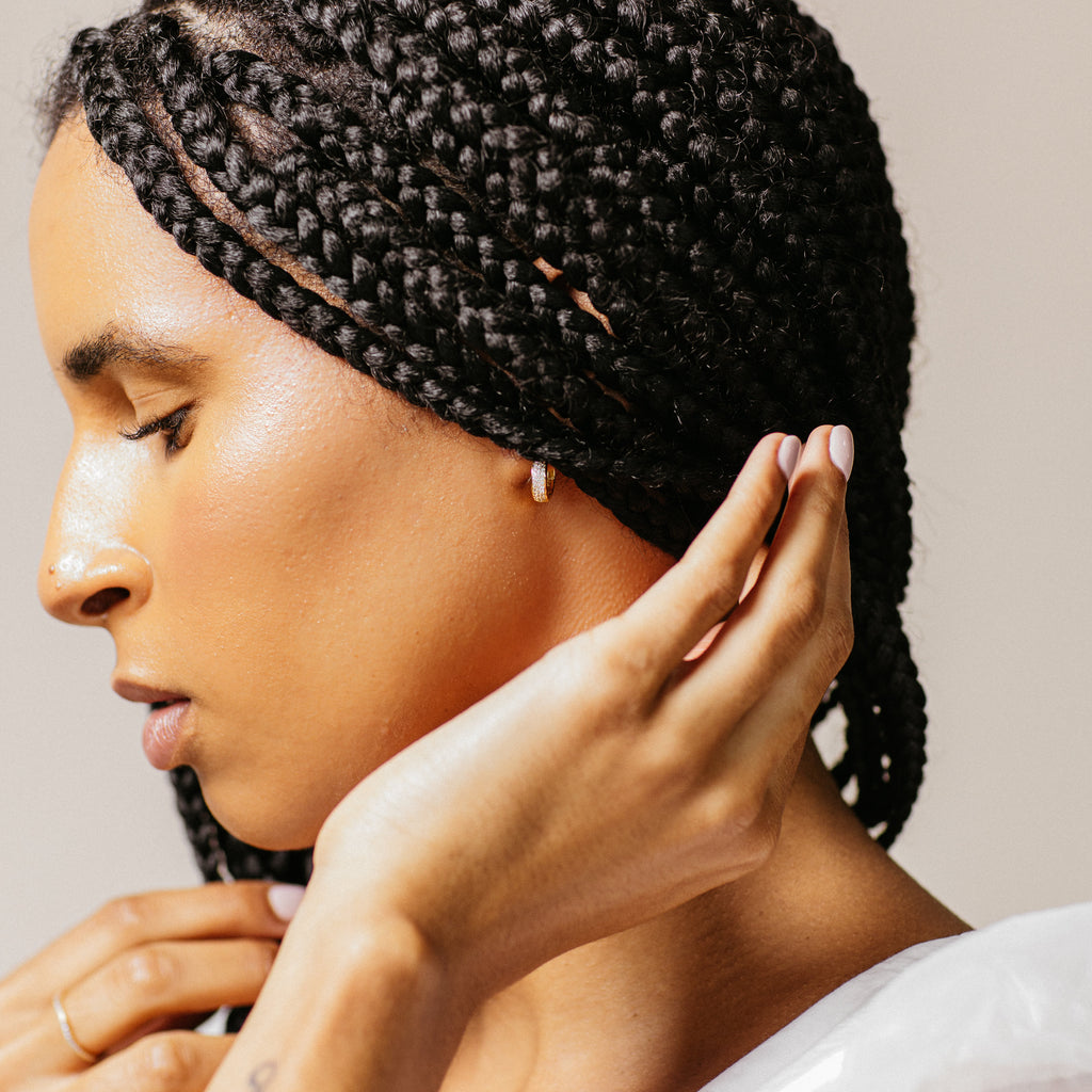 A woman with braided hair touches her ear, highlighting the sparkle of Pave Diamond Huggies in soft lighting—these earrings are must-have jewelry box essentials.