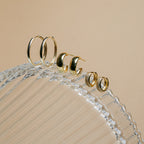 Pave Diamond Huggies are displayed alongside two other gold hoops on the edge of a clear, rippled glass dish set against a beige backdrop—a true jewelry box essential.