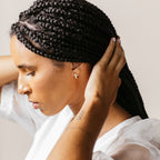 Woman with braided hair in profile, wearing a white top and touching her head with both hands, showcases Pave Diamond Huggies—timeless jewelry box essentials.
