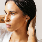 A person with braided hair wearing a white top touches the back of their head, showcasing the Pave Diamond Huggies—sparkling diamond-encrusted hoops that are a must-have jewelry box essential.