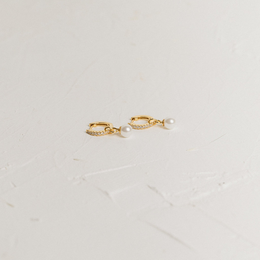 Mae Huggies: Gold hoop earrings accented with pearls and small crystals are displayed on a textured white surface—perfect for those who love timeless pearl huggies.