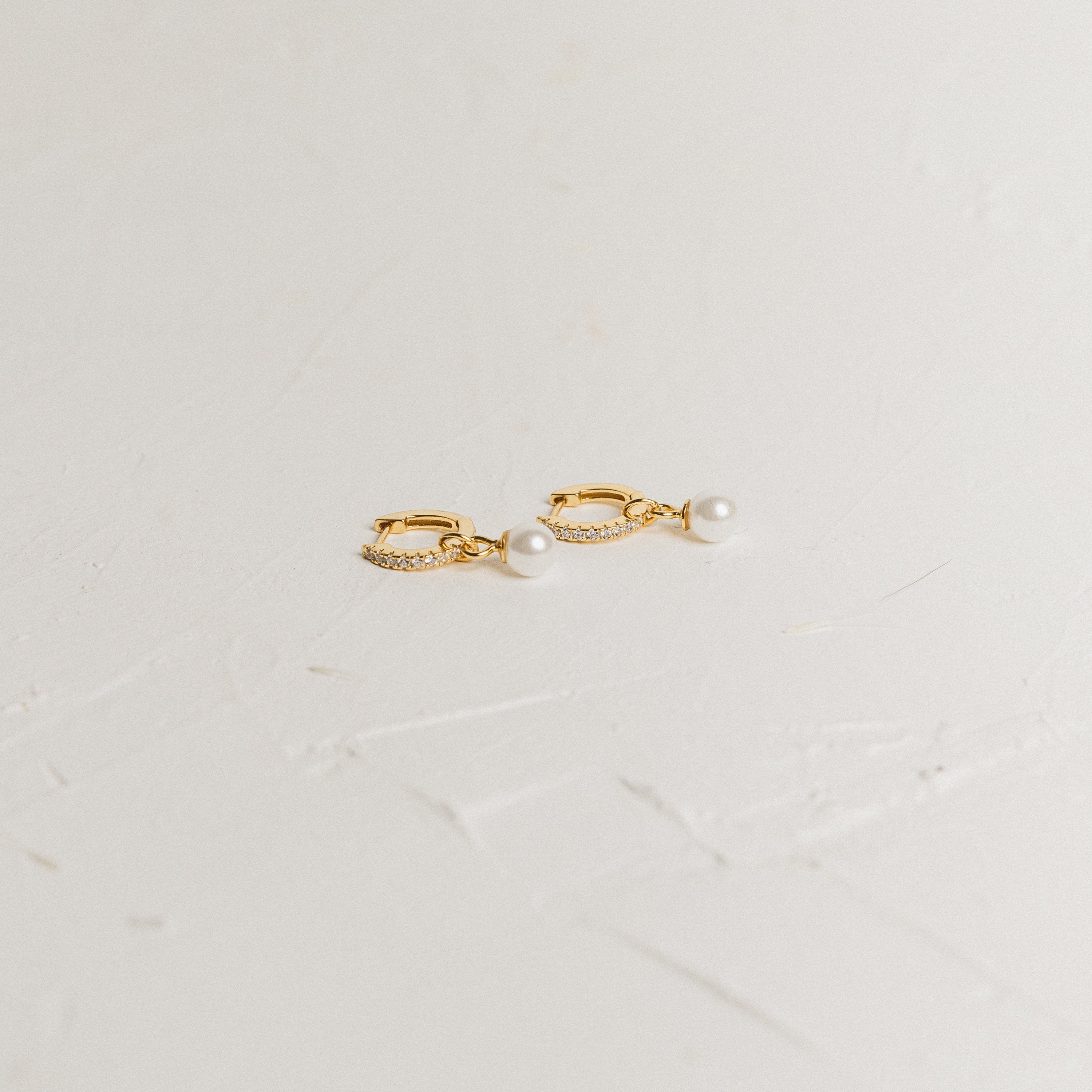 Mae Huggies: Gold hoop earrings accented with pearls and small crystals are displayed on a textured white surface—perfect for those who love timeless pearl huggies.