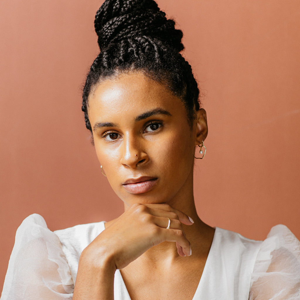 A woman with braided hair in a bun, dressed in a white top, models the Mae Huggies earrings while posing with her hand under her chin against a peach background.