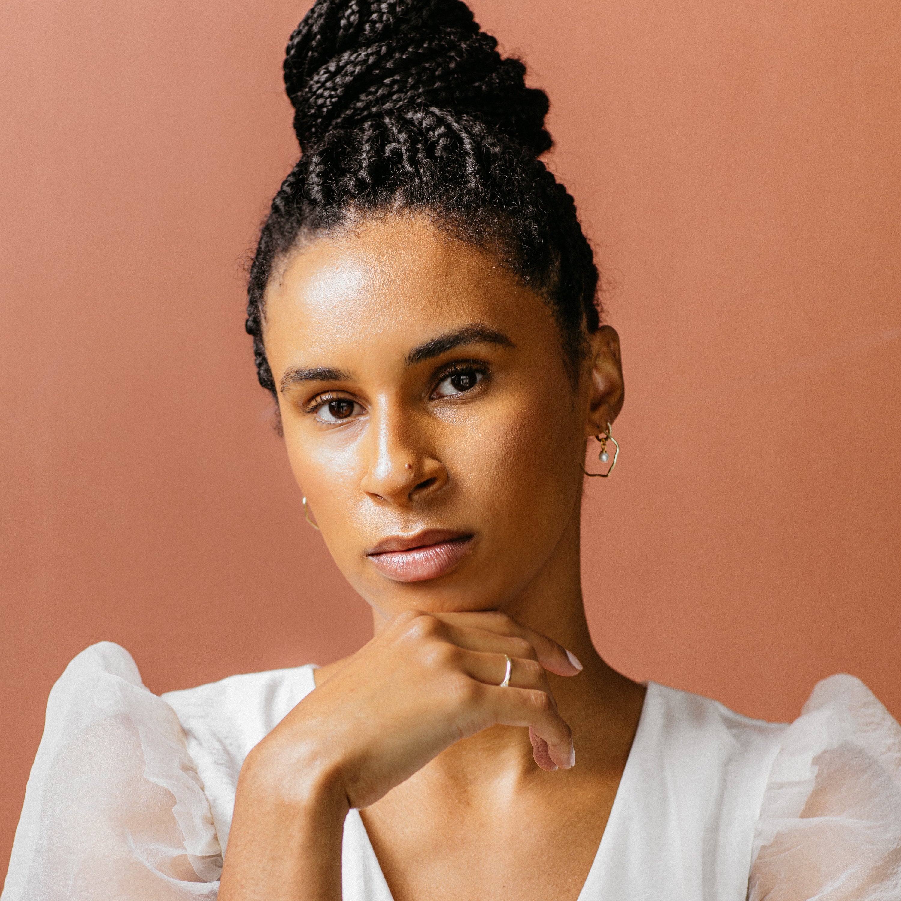 A woman with braided hair in a bun, dressed in a white top, models the Mae Huggies earrings while posing with her hand under her chin against a peach background.