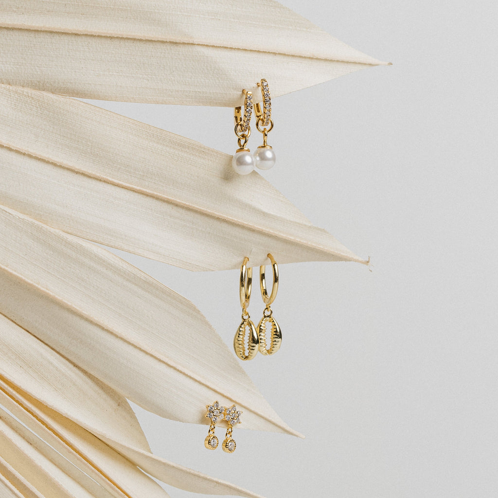 Three pairs of elegant earrings, including the Mae Huggies, shell designs, and diamond-encrusted hoop studs, displayed on a dried palm leaf.