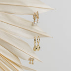 Three pairs of elegant earrings, including the Mae Huggies, shell designs, and diamond-encrusted hoop studs, displayed on a dried palm leaf.