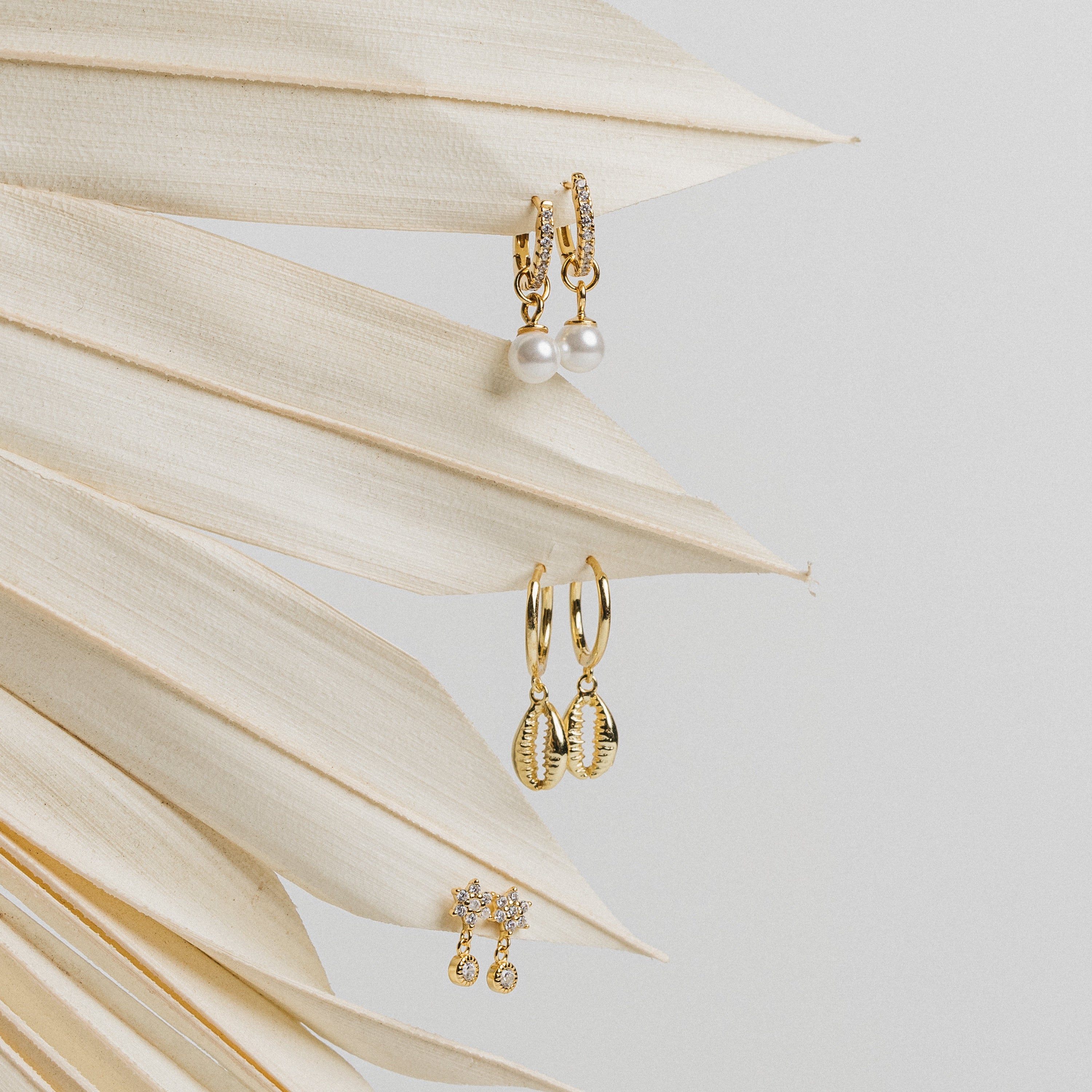 Three pairs of elegant earrings, including the Mae Huggies, shell designs, and diamond-encrusted hoop studs, displayed on a dried palm leaf.