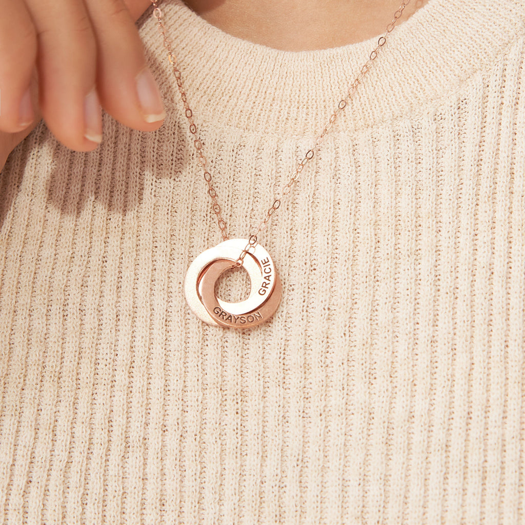 Hand holding the Interlocking Family Necklace in rose gold, featuring a circular custom-engraved family pendant with GRAYSON and GRACE, over a beige sweater.