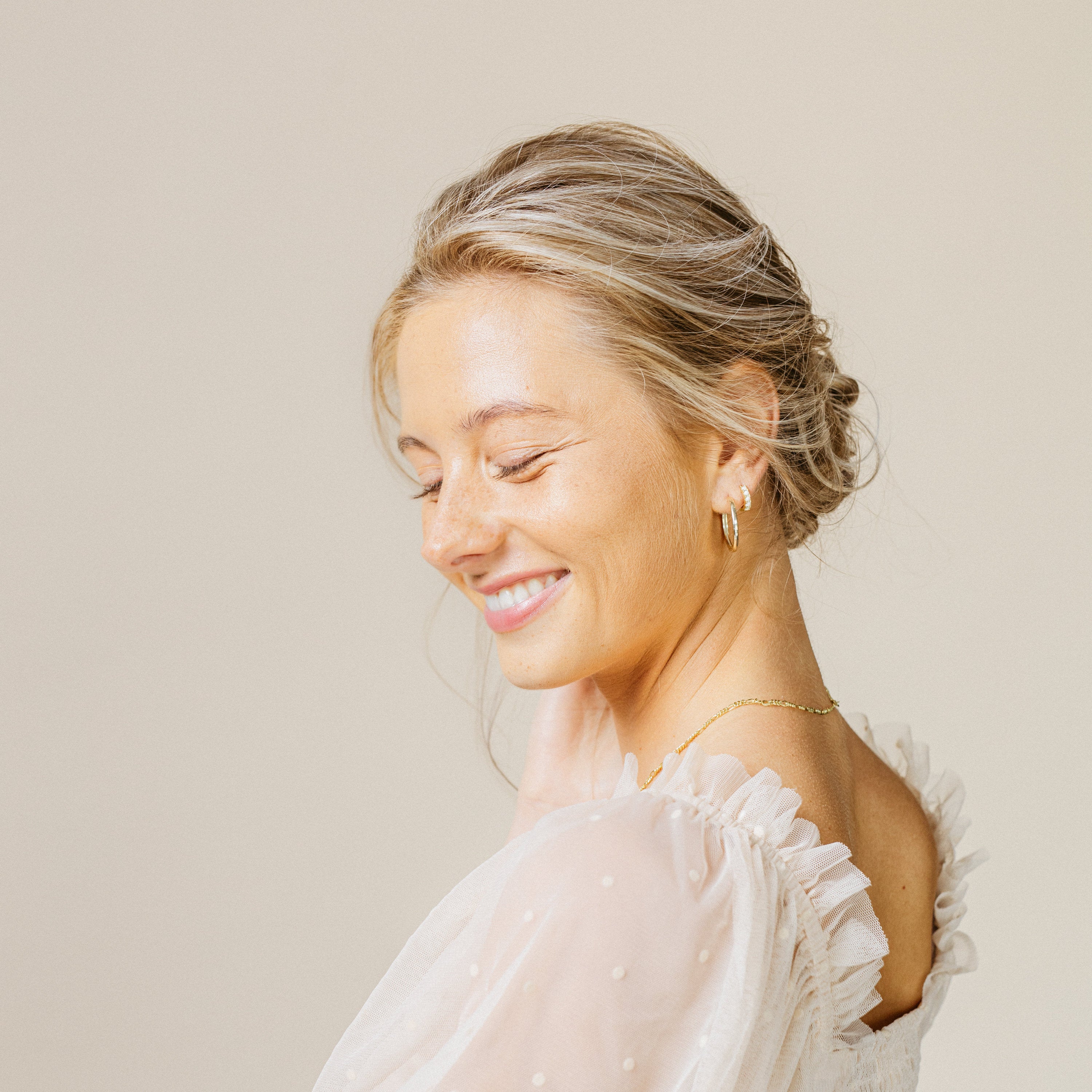 Smiling blonde woman with hair in a loose updo wears minimal earrings—Opal Huggies—and a white ruffled top as she looks down.