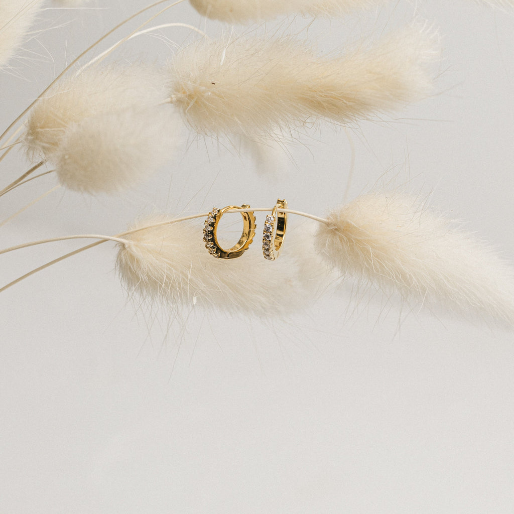 The Coco Pave Huggies in 18K Gold, featuring delicate crystals, hang on soft white dried grasses against a pale backdrop, showcasing their elegant and sparkling design.