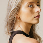 Woman with long blonde hair wearing Coco Pave Huggies in 18K Gold and a black top, looking over her shoulder.