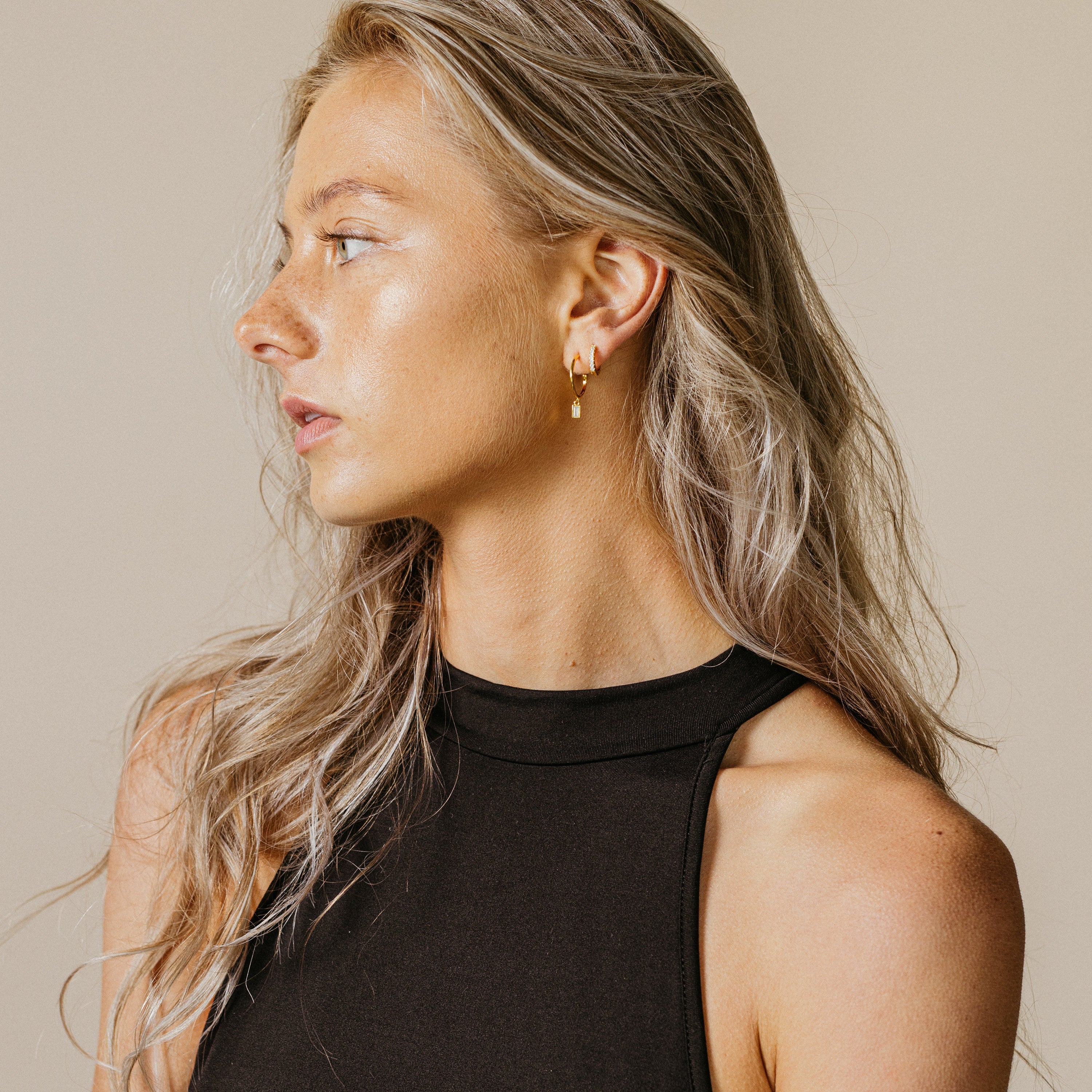 Woman with long blonde hair wearing a black sleeveless top and shimmering Elle Huggies, looking to the left.