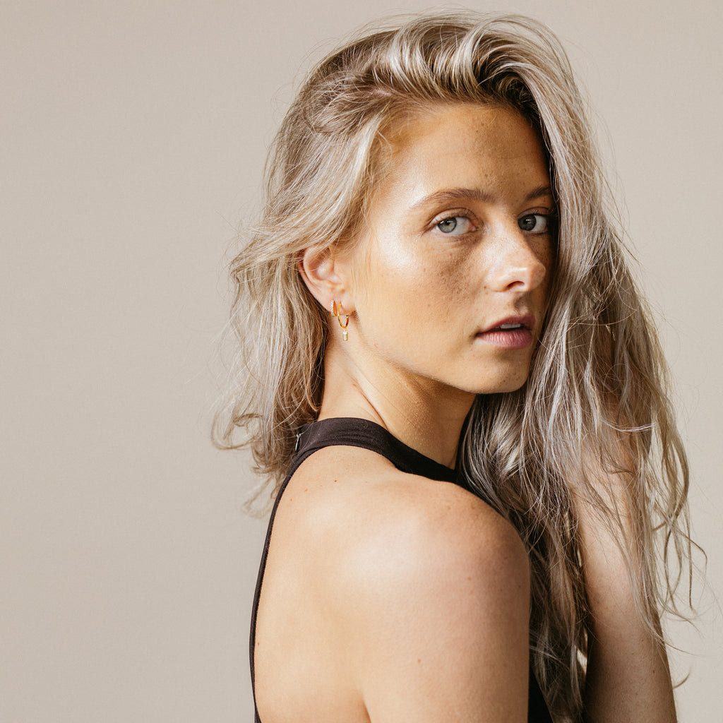 A woman with long, wavy blonde hair poses against a neutral background, elegantly showcasing sparkling Elle Huggies.