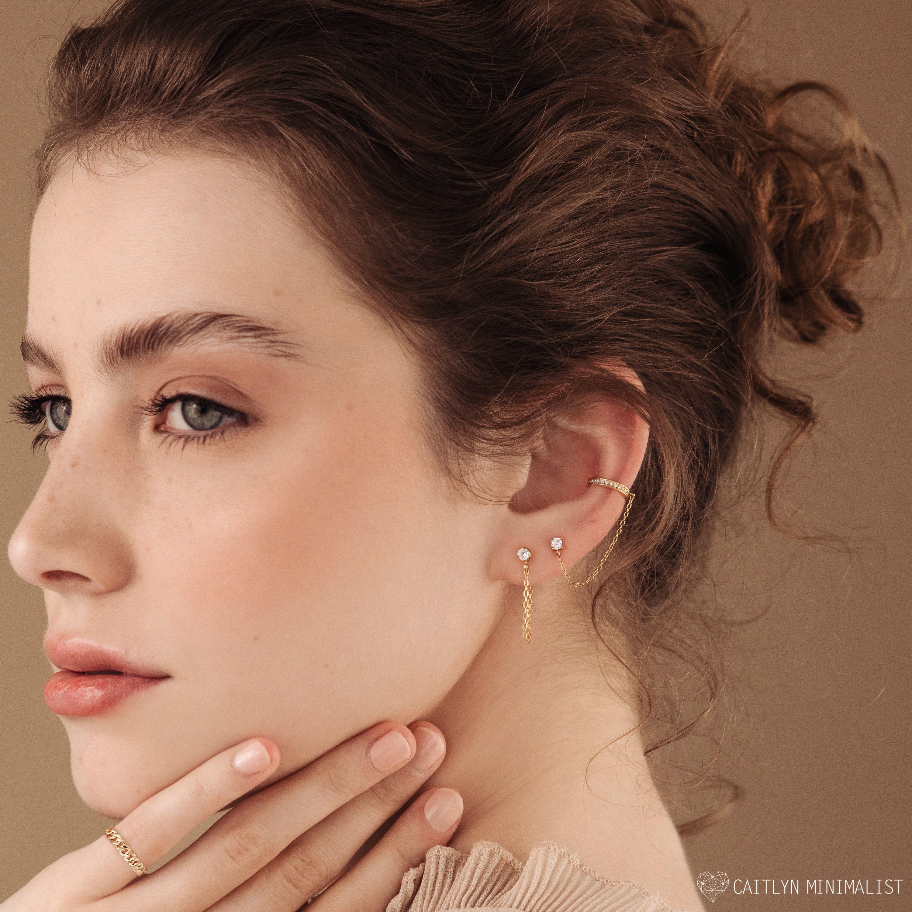 Woman with curly brown hair wears Birthstone Chain Studs and a gold ring, gently touching her chin—showcasing versatile jewelry perfect for any occasion.