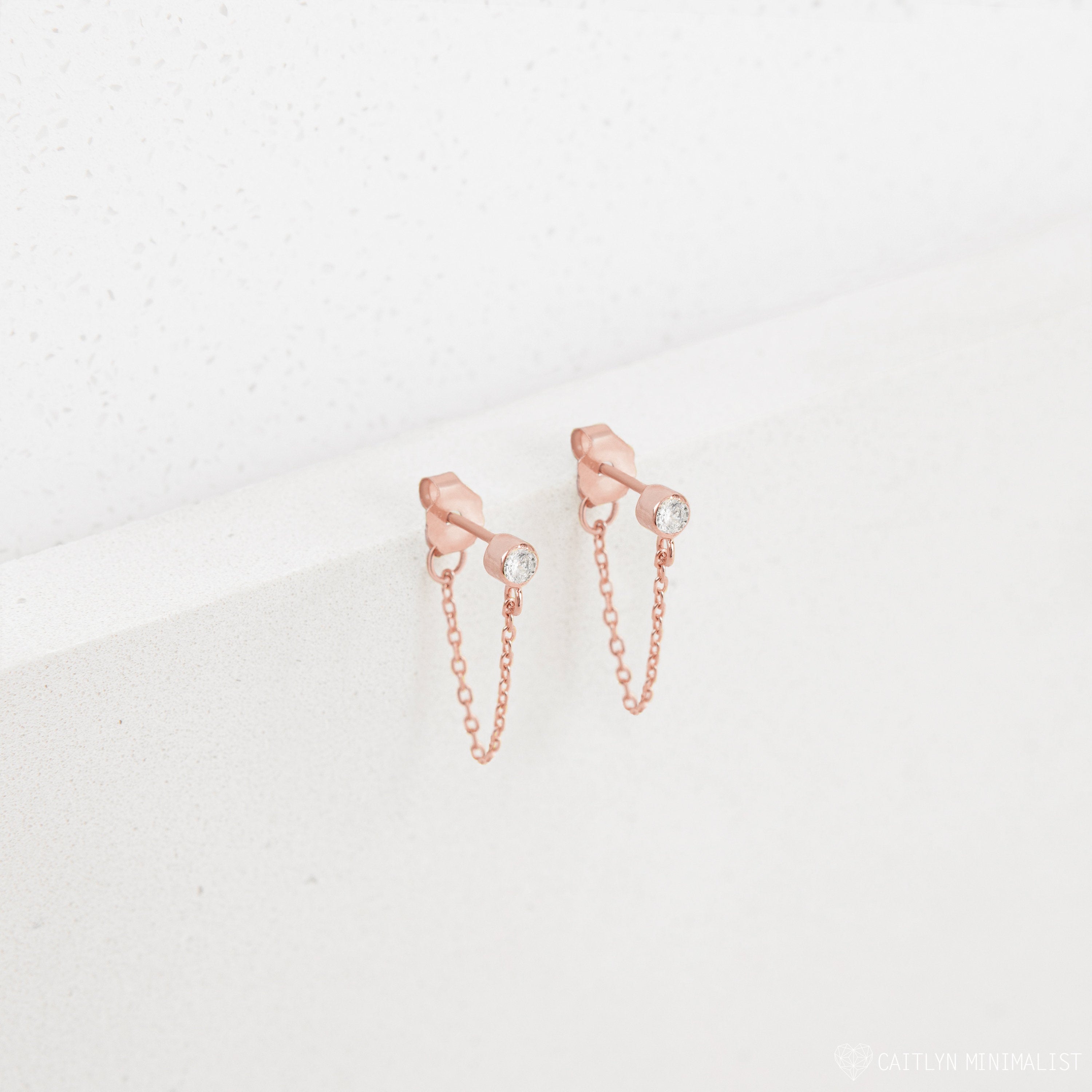 Birthstone Chain Studs in rose gold feature clear gemstones and a delicate chain, shown on a white textured background—versatile earrings ideal for any occasion.