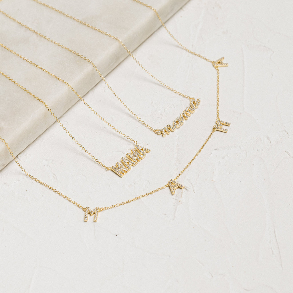The Pave Mama Script Necklace features gold letter charms that spell “MAMA” and “mama” on a light textured surface—perfect Motherhood Jewelry and an ideal Gift for Mom.