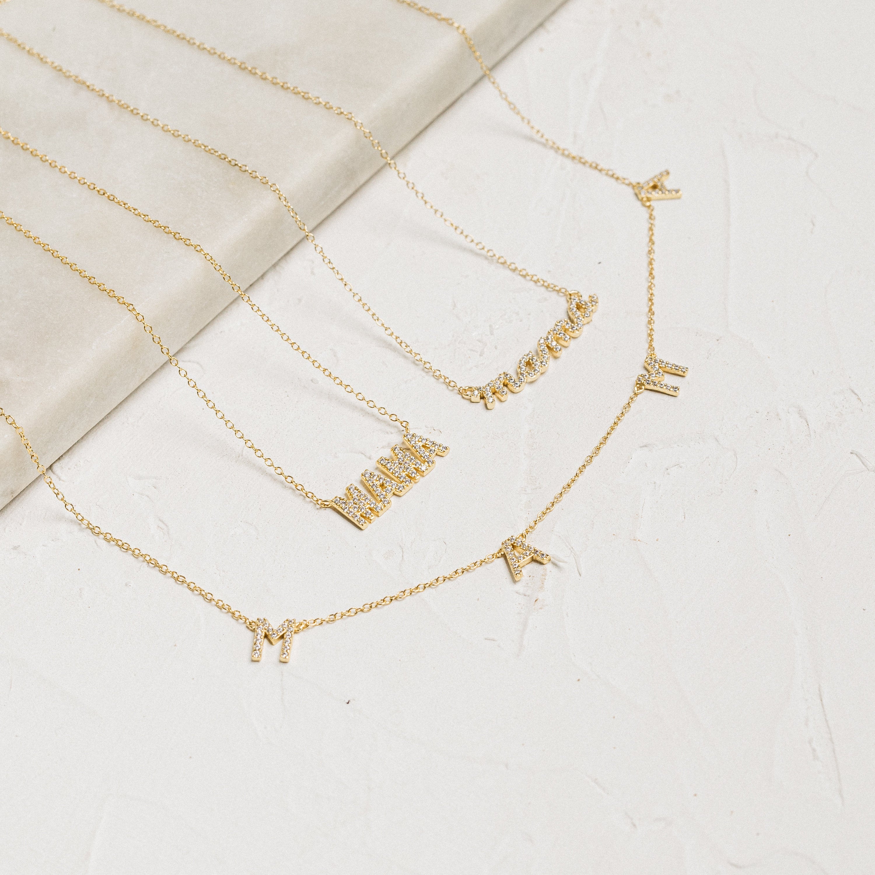 The Pave Mama Script Necklace features gold letter charms that spell “MAMA” and “mama” on a light textured surface—perfect Motherhood Jewelry and an ideal Gift for Mom.