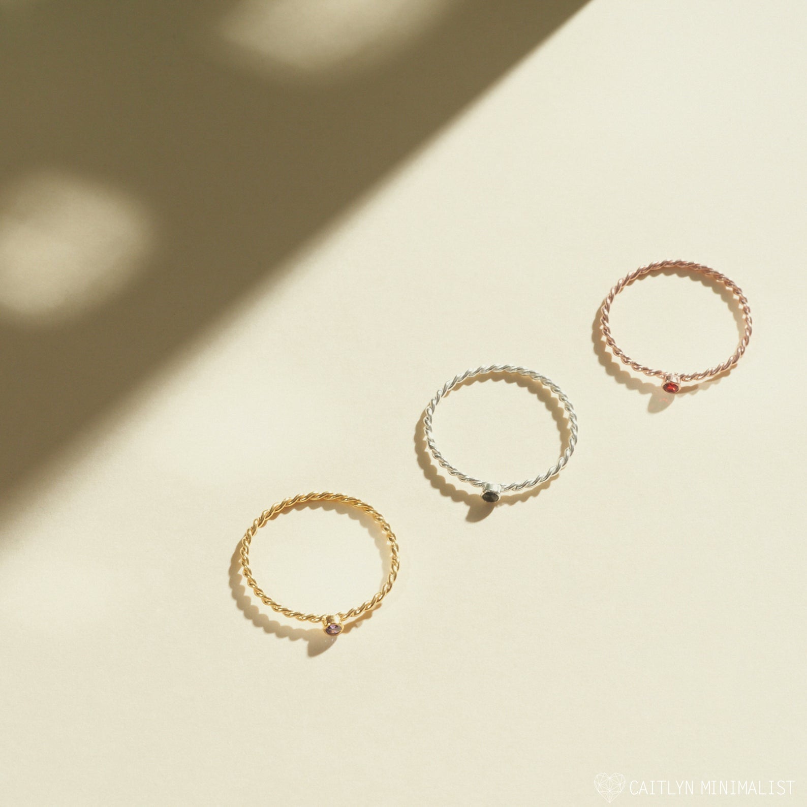 The Twisted Birthstone Ring, shown in gold, silver, and rose gold on a beige surface with soft shadows, is perfect for stacking.