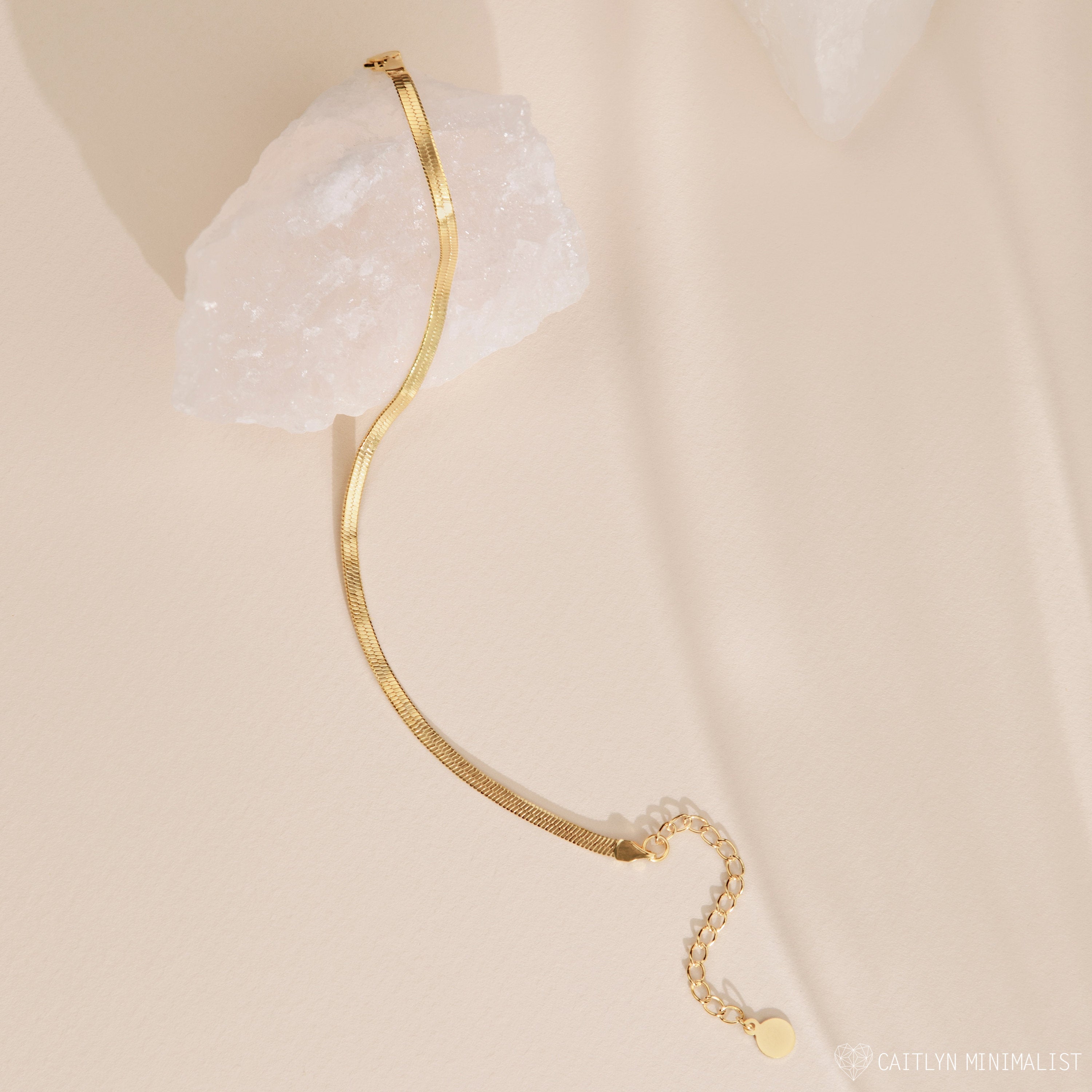 A Herringbone Anklet draped over a white crystal on a beige surface, highlighting the polished ankle jewelry's effortless elegance.