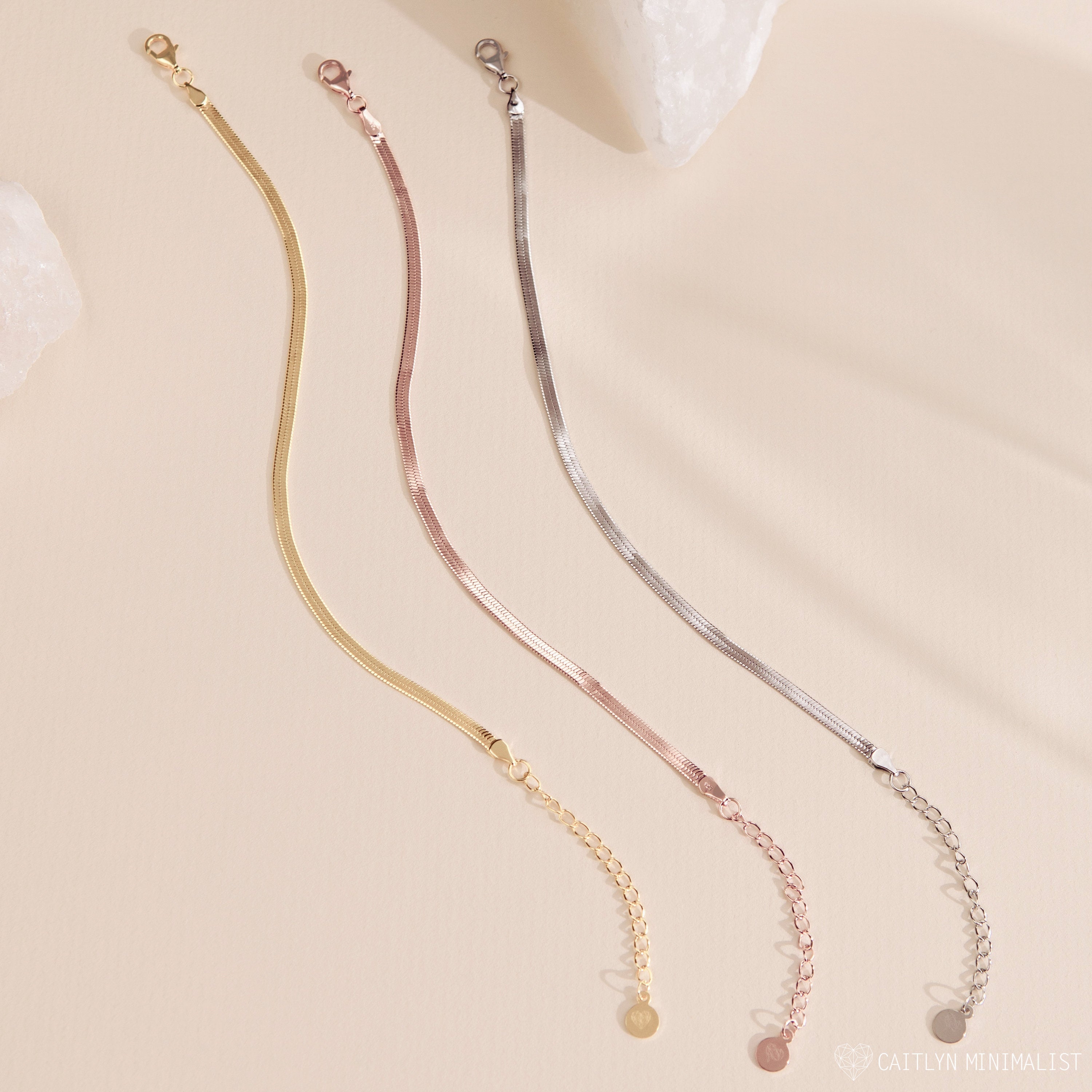 Three minimalist chain bracelets in gold, rose gold, and silver are displayed on a light beige background, perfectly complementing any polished ankle jewelry or pairing seamlessly with the Herringbone Anklet.