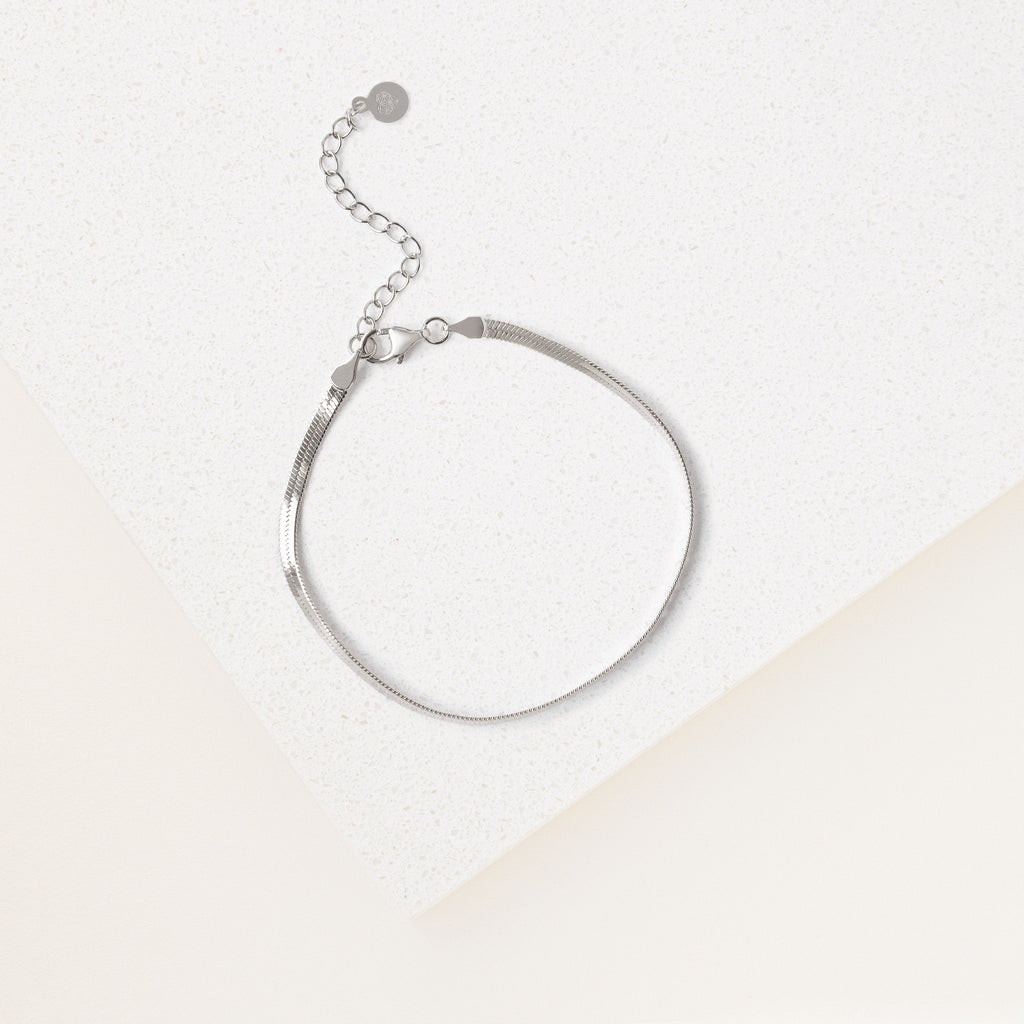 The Herringbone Bracelet features a contemporary silver design and adjustable clasp, showcased on a white textured surface beneath soft lighting.