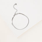 The Herringbone Bracelet features a contemporary silver design and adjustable clasp, showcased on a white textured surface beneath soft lighting.