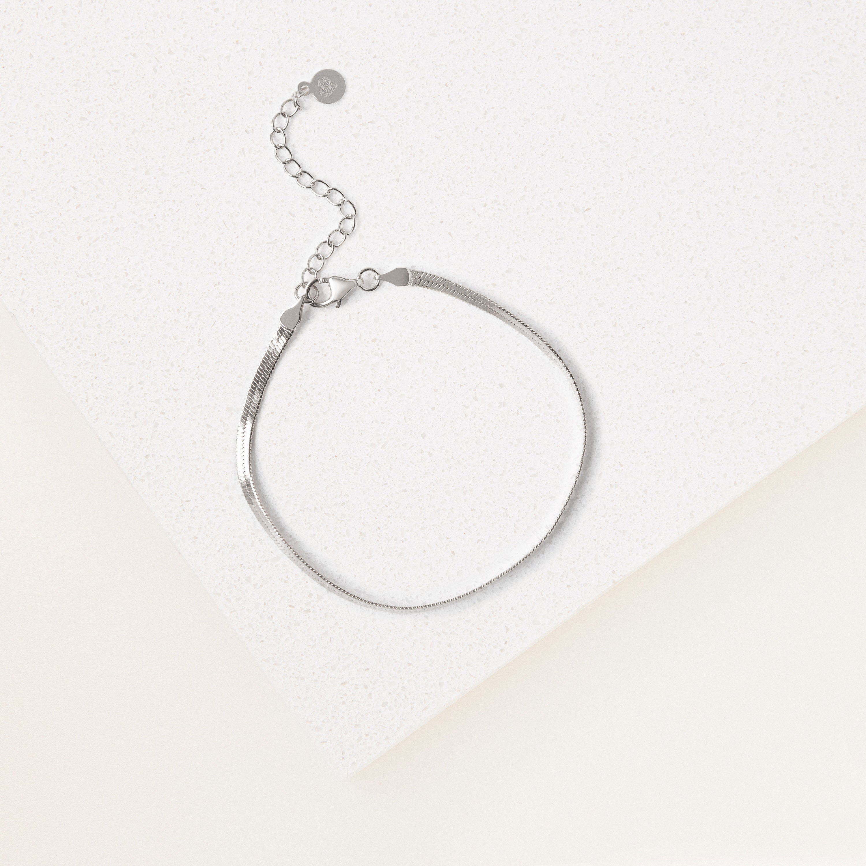 The Herringbone Bracelet features a contemporary silver design and adjustable clasp, showcased on a white textured surface beneath soft lighting.