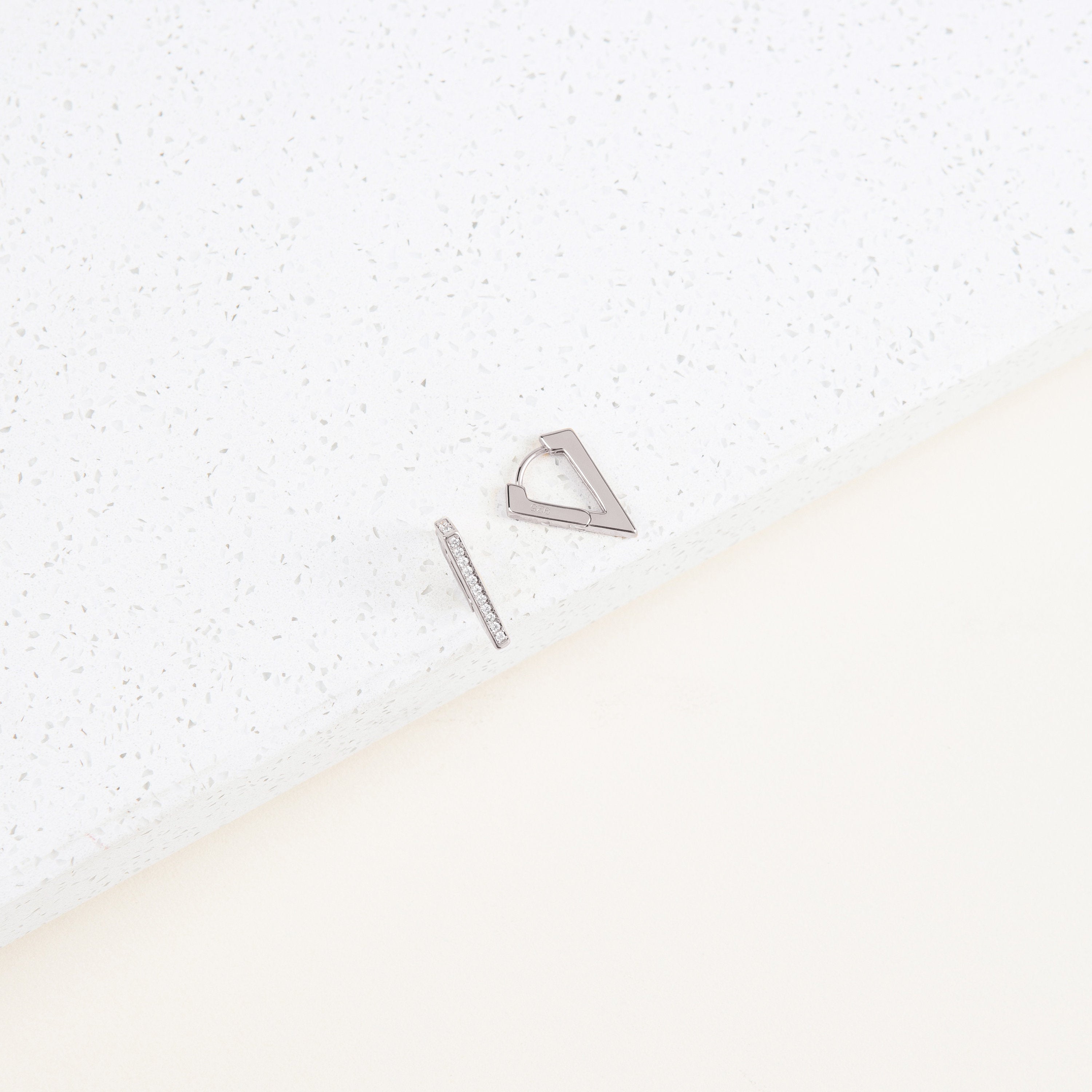 The Ezra Triangle Earrings feature two minimalist silver geometric designs—one with a pavé-textured bar and the other a sleek triangle—resting on a white speckled surface.