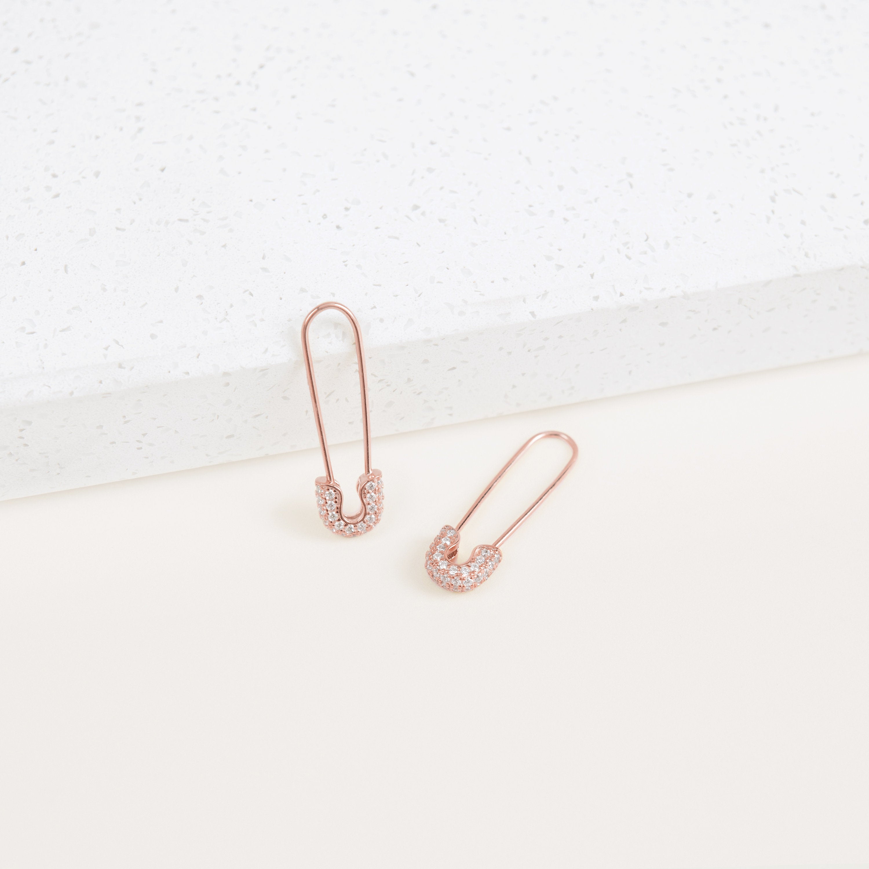 Two Pave Safety Pin Earrings in rose gold, featuring sparkling rhinestones, are displayed on a white textured surface.