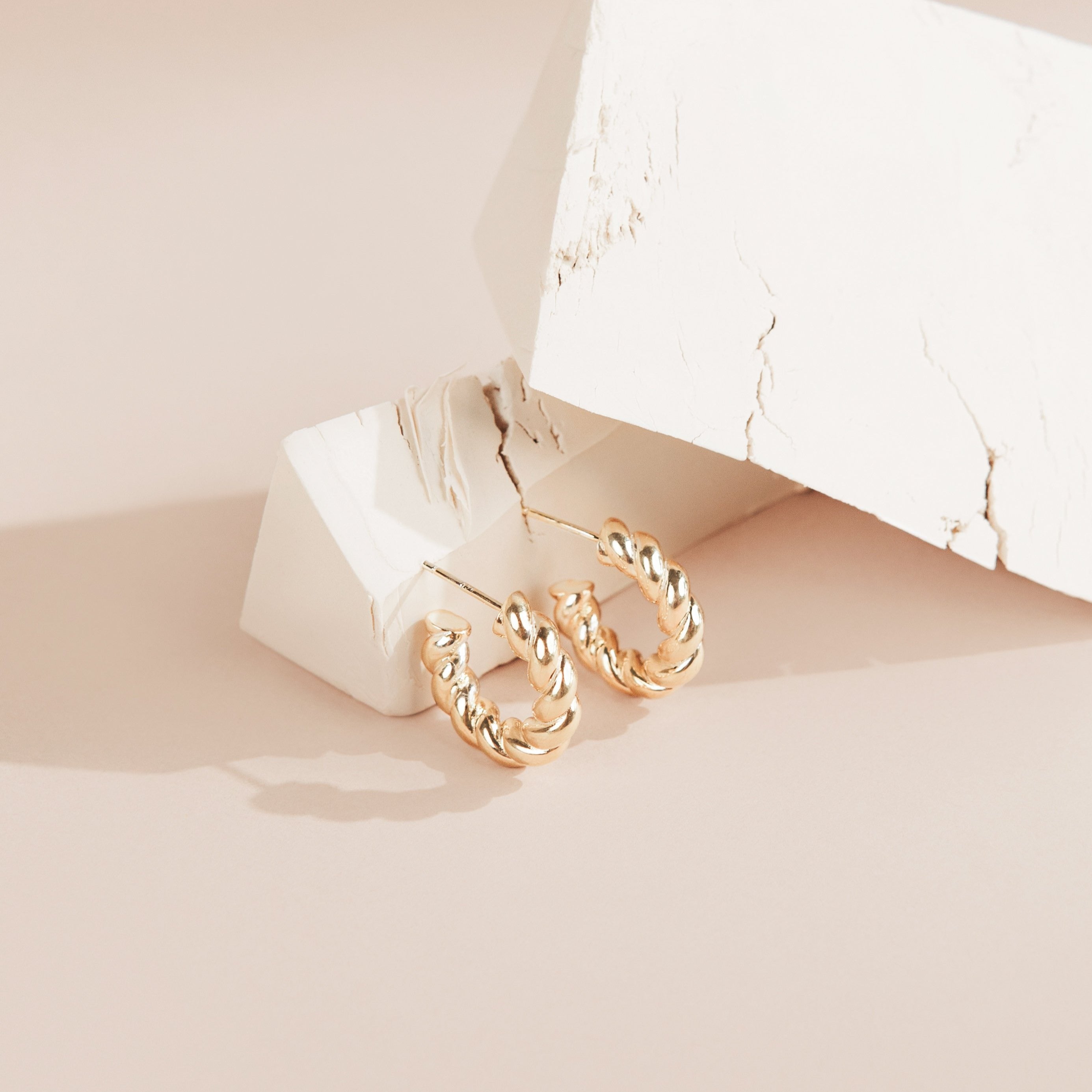 Remi Twist Hoops showcased on white textured blocks atop a soft beige surface—perfect for minimal jewelry lovers.